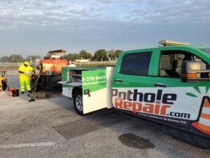PotholeRepair.com service truck and crew member preparing thermal asphalt repair equipment in a commercial parking lot at sunrise
