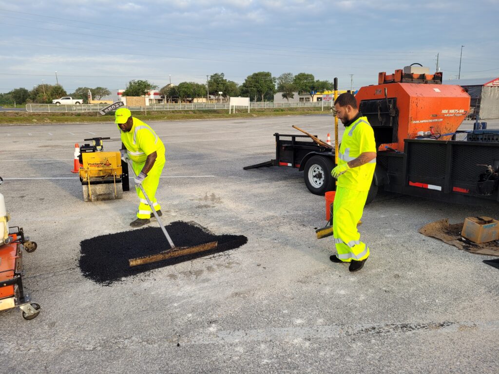 PotholeRepair.com crew working hot asphalt into a square-cut pothole repair in a commercial parking lot, with a compactor and the company heated material trailer on site