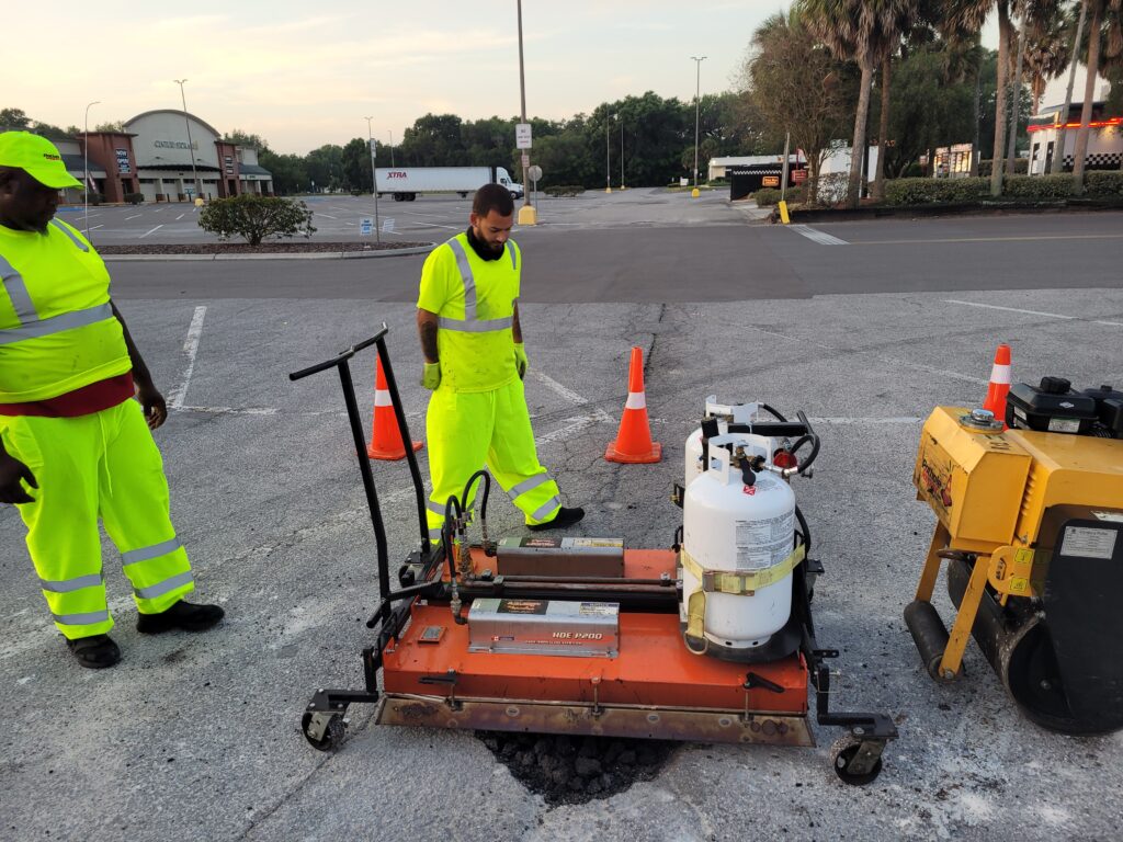 Two PotholeRepair.com crew members in hi-vis running an infrared heater over a parking lot pothole repair, with the roller and coned work zone in a retail center lot at dawn