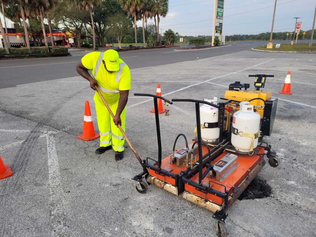PotholeRepair.com technician using a propane infrared asphalt heater to repair a pothole in a commercial retail parking lot, with safety cones set around the work area