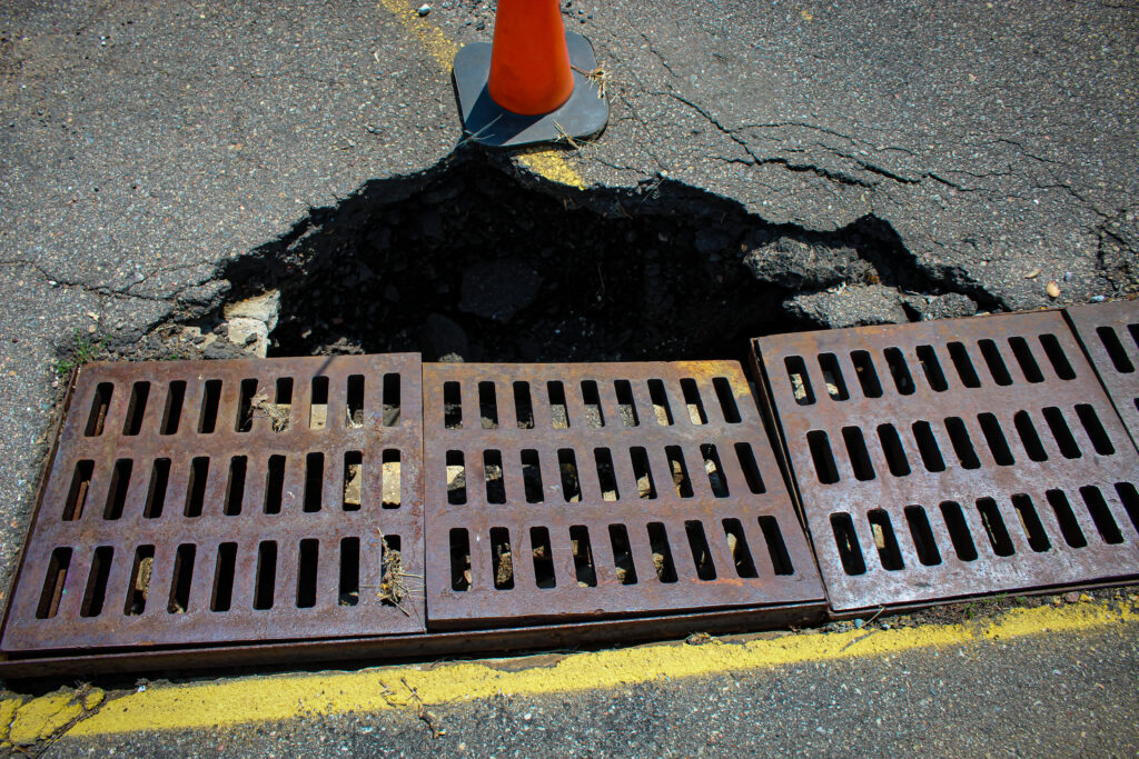Coned pothole with collapsed asphalt beside storm drain grates in a commercial parking lot, the drainage-driven base failure a professional pavement evaluation documents