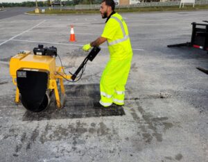 PotholeRepair.com crew member in hi-vis rolling a fresh thermal pothole repair on a commercial parking lot at dawn, compacting it tight while the asphalt is still hot