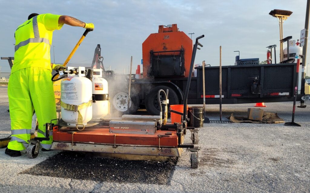 PotholeRepair.com technician lifting the infrared heater off a freshly heated pothole repair in a commercial parking lot, steam rising, with the company heated material trailer behind